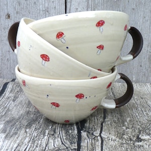 Könnte beinhalten: Drei gestapelte Keramikschalen mit cremefarbener Glasur. Jede Schale ist mit roten Pilzillustrationen und dunkelbraunen Griffen verziert. Die Schalen stehen auf einer Holzoberfläche.