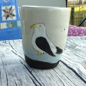 Könnte beinhalten: Eine Keramiktasse mit handgemaltem Design einer Möwe mit schwarzen und weißen Markierungen und gelben Füßen. Die Tasse hat eine hellbraune Glasur mit blauen und weißen Akzenten.