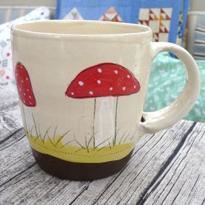 Könnte beinhalten: Cremefarbene Keramiktasse mit handgemalten roten Pilzen mit weißen Punkten. Die Pilze sind vor Gras und einem braunen Sockel platziert. Die Tasse hat einen großen Henkel und ist für Heißgetränke geeignet.