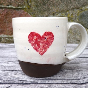 Könnte beinhalten: Weiße Keramiktasse mit rotem Herz-Design und braunem Rand. Die Tasse hat eine gesprenkelte Glasur und einen Henkel.