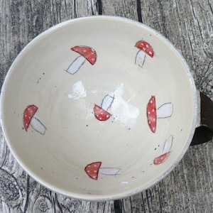 Könnte beinhalten: Cremefarbene Keramikschale mit dunkelbraunem Griff, verziert mit roten und weißen Pilzillustrationen. Die Schale hat einen breiten Rand und ist mit kleinen schwarzen Punkten versehen. Ein skurriles Küchenutensil mit Naturthema.
