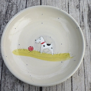 Könnte beinhalten: Weißer Keramikteller mit handgemalter Illustration eines Hundes und eines roten Balls. Der Hund ist weiß mit schwarzen Flecken und rotem Halsband, steht auf gelbem Gras. Der Teller hat ein schlichtes, rustikales Design.