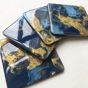 Puede incluir: Juego de cinco posavasos cuadrados con acabado brillante. Los posavasos presentan un diseño veteado en azul marino y azul claro con detalles de purpurina dorada. Los posavasos están dispuestos en un patrón ligeramente superpuesto.