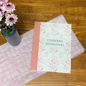 Op de afbeelding: Een notitieboek met de tekst "CUADERNO DEVOCIONAL" ligt op roze vloeipapier. Het notitieboek heeft een roze rug en een witte kaft met een roze bloemenpatroon. Links staat een vaas met paarse bloemen.