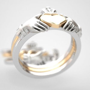 May include: A silver and gold Claddagh ring with two hands clasped around a heart. The heart is gold and has a crown on top.