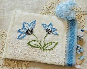 Pode incluir: Uma toalha de chá de linho branco com flores bordadas azuis e uma borda azul e branca. A toalha é decorada com um delicado padrão de renda.