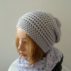 K&ouml;nnte beinhalten: Eine hellgraue, geh&auml;kelte Beanie-M&uuml;tze mit offenem Webmuster. Die M&uuml;tze wird auf einem Schaufensterkopf pr&auml;sentiert, kombiniert mit einem floralen Schal. Die Textur und Farbe der M&uuml;tze deuten auf ein handgefertigtes, bequemes Accessoire f&uuml;r k&uuml;hleres Wetter hin.