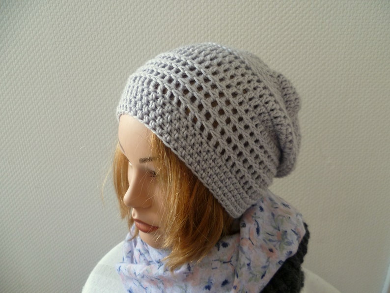 K&ouml;nnte beinhalten: Eine hellgraue, geh&auml;kelte Beanie-M&uuml;tze mit offenem Webmuster. Die M&uuml;tze wird auf einem Schaufensterkopf pr&auml;sentiert, kombiniert mit einem floralen Schal. Die Textur und Farbe der M&uuml;tze deuten auf ein handgefertigtes, bequemes Accessoire f&uuml;r k&uuml;hleres Wetter hin.