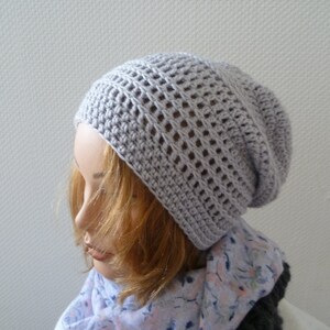 K&ouml;nnte beinhalten: Hellgraue, geh&auml;kelte Beanie-M&uuml;tze mit offenem Webmuster. Die M&uuml;tze wird auf einem Schaufensterkopf mit blondem Haar und einem floralen Schal getragen. Die M&uuml;tze hat eine weiche, neutrale Farbe.