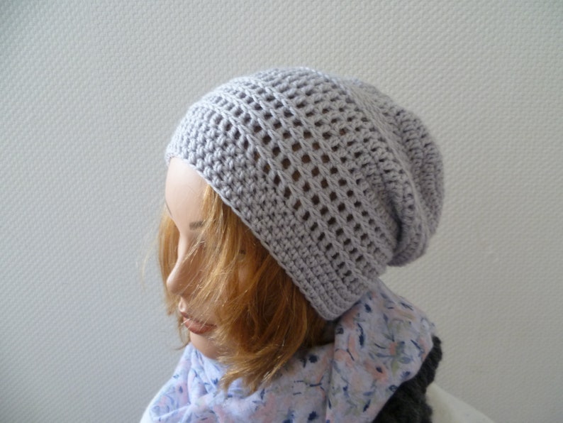 K&ouml;nnte beinhalten: Hellgraue, geh&auml;kelte Beanie-M&uuml;tze mit offenem Webmuster. Die M&uuml;tze wird auf einem Schaufensterkopf mit blondem Haar und einem floralen Schal getragen. Die M&uuml;tze hat eine weiche, neutrale Farbe.