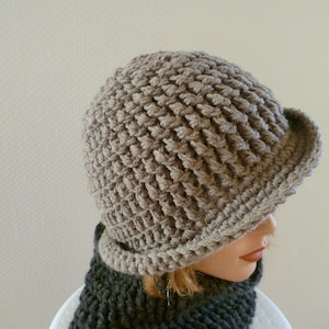 Könnte beinhalten: Gehäkelter Hut mit kleinem Schirm in neutraler Beigetönung. Der Hut hat ein strukturiertes, gewebtes Muster. Ein dunkelgrauer Schal ist unter dem Hut zu sehen.