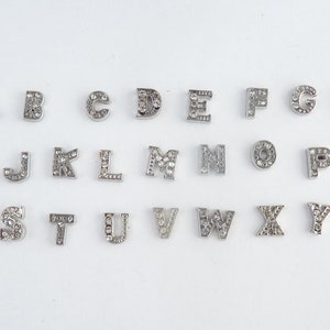 Può includere: Un set di 26 ciondoli argentati, ognuno con una lettera dell'alfabeto adornata con strass trasparenti.