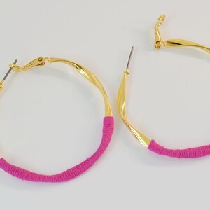 Può includere: Un paio di orecchini a cerchio dorati con un filo rosa avvolto attorno alla metà inferiore di ogni cerchio.