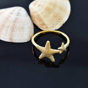 Puede incluir: Un anillo de oro con dos diseños de estrellas de mar. El anillo está sobre un fondo negro con dos conchas.