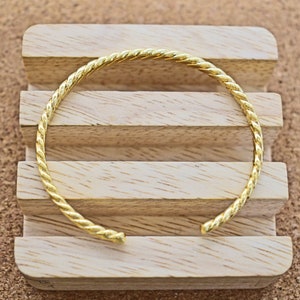 Könnte beinhalten: Ein goldfarbenes, gedrehtes Armkettchen auf einem Holzständer.
