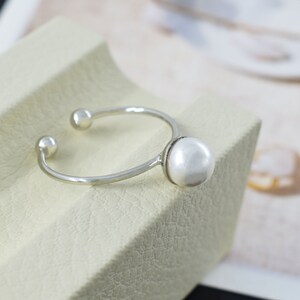 Può includere: Un anello d'argento con un grande perla bianca sul davanti. L'anello è aperto sul retro e ha due piccole sfere d'argento sui lati.