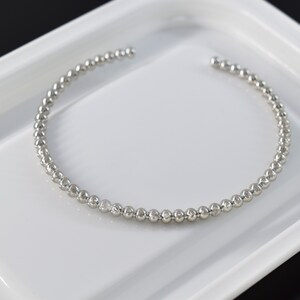 Puede incluir: Una pulsera de cuentas plateada con un diseño abierto. La pulsera está compuesta por pequeñas cuentas redondas y descansa sobre una superficie blanca. El fondo es negro.