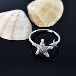 May include: A silver ring with two starfish designs. The ring is on a black background with two seashells.