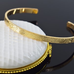 Könnte beinhalten: Ein goldfarbenes Armreif-Armband mit strukturierter Oberfläche. Das Armband ist offen und hat ein schlichtes, minimalistisches Design.
