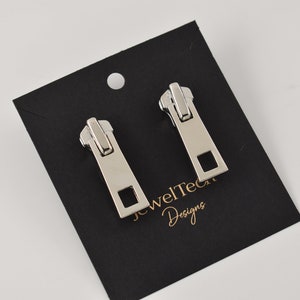 Op de afbeelding: Een paar zilveren rits-oorbellen op een zwarte kaart met de tekst "JewelTec Designs".