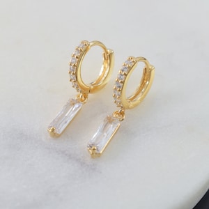 May include: Gold hoop earrings with a dangling rectangular clear crystal. The hoops are lined with small clear crystals.