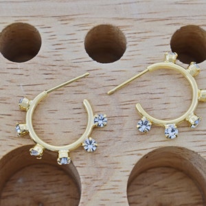 May include: A pair of gold hoop earrings with clear crystal accents. The earrings are on a wooden surface.