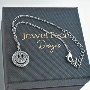 May include: A silver chain bracelet with a sparkly smiley face charm. The bracelet is in a black box with the text "Jewel Tech Designs" on it.