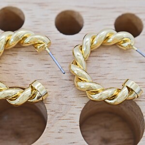 May include: A pair of gold-toned hoop earrings with a twisted rope design. The earrings are displayed on a wooden surface.