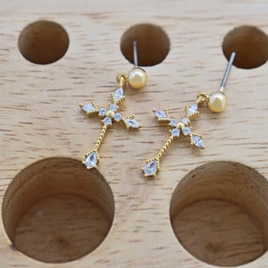May include: A pair of gold earrings with a cross design and clear crystal accents. The earrings have a pearl-like drop at the bottom.
