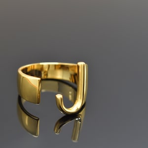 May include: A gold-colored ring with the letter "J" in a bold, modern font. The ring is open and adjustable.