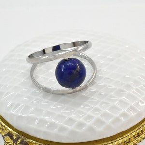 Può includere: Un anello d'argento con un pendente a forma di gemma sferica blu scuro. L'anello ha un design unico con due fasce curve. La gemma è di un blu intenso, su sfondo bianco.