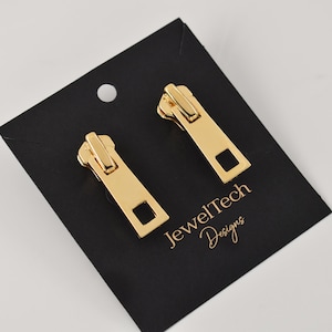May include: A pair of gold zipper pull earrings on a black card with the text "JewelTech Designs".