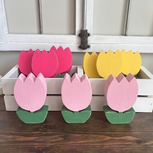 May include: A set of wooden tulip flower decorations in pink, yellow, and red. The tulips are painted in solid colors and have green leaves. The tulips are arranged in a white wooden crate.