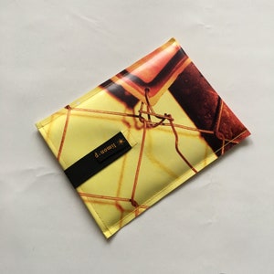 Könnte beinhalten: Eine gelbe Tasche mit einem schwarzen Riemen und dem Text "limon p." in goldenen Buchstaben. Die Tasche hat ein Muster aus einem Maschendrahtzaun mit einem rostigen Metalltor.
