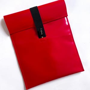 Könnte beinhalten: Eine leuchtend rote, rechteckige Tasche mit glänzender Oberfläche. Ein schwarzes Gummiband sichert die gefaltete Oberseite. Auf dem Band steht der Text "limón-t". Die Tasche dient wahrscheinlich zum Tragen von Gegenständen.