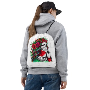 May include: White drawstring bag with a colorful illustration of a woman wearing a headscarf and a floral crown. The woman's face is partially covered by the headscarf, and she is looking to the side. The background is white with splashes of color.