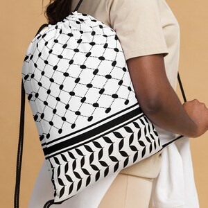 May include: White drawstring backpack with a black geometric pattern. The design features a repeating pattern of black ovals connected by dotted lines, with a border of black leaves. The bag has black drawstrings.