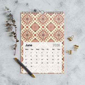以下が含まれることがあります： クリーム色の背景に赤とクリーム色の幾何学模様が繰り返される、2026年6月の壁掛けカレンダー。カレンダーには金属製のスパイラルバインディングとペンが付いています。