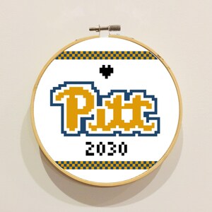 May include: A circular wooden embroidery hoop containing white fabric with a cross-stitch design. The design features the text "Pitt" in yellow with a blue outline, a small black pixelated heart, the number "2030" in black, and blue and yellow checkered borders.