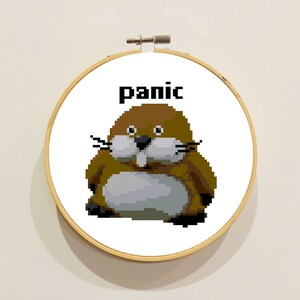 May include: A circular wooden embroidery hoop framing a white fabric piece with a pixel art design of a brown and gray groundhog. Above the groundhog, the word "panic" is stitched in black pixelated letters. The hoop has a metal tension screw.
