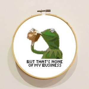 Puede incluir: Un aro de bordado blanco con una rana verde bebiendo de una taza. La rana es verde con un ojo blanco y el texto "BUT THAT'S NONE OF MY BUSINESS" está impreso debajo de la rana.