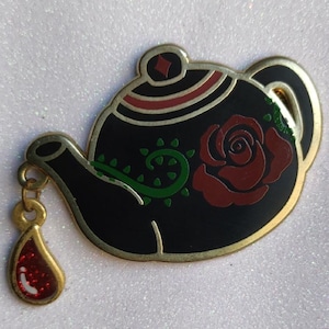 Puede incluir: Un broche de tetera negra con borde dorado y diseño de rosa roja. El broche tiene un dije de lágrima dorada con purpurina roja.  Vides verdes y detalles rojos decoran la tetera.
