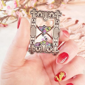 May include: A silver-colored hourglass pin with a clear center filled with colorful floral and character designs. The pin has a rectangular frame with decorative elements at the top, bottom, and sides. The pin is held in a hand with red nail polish.