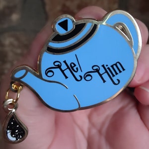 May include: A light blue enamel pin in the shape of a teapot with gold trim. The pin has the words "He/Him" in black script. A small charm in the shape of a teardrop hangs from the teapot.