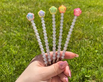 May include: Five decorative pearl-beaded hair pins. Each pin features a different pastel-colored star-shaped charm. The charms are pink, yellow, blue, green, and orange. The pins are held against a backdrop of green grass.