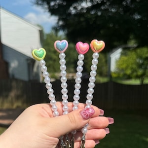 May include: Four beaded hair clips with heart-shaped toppers in pastel colors: yellow, blue, pink, and gold. Each clip has a string of white pearls leading to a silver metal clip. The background is a green lawn and trees.