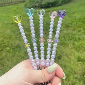 May include: Five hair clips with pearl beads and heart-shaped crystal toppers in yellow, blue, clear, pink, and purple. Each clip has a small flower accent. The clips are held in a hand against a blurred green background.