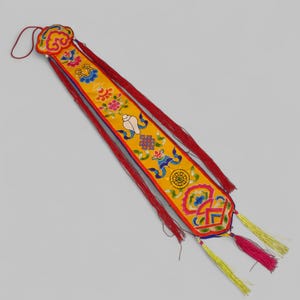 Könnte beinhalten: Ein langes, gelbes Textil mit roten Quasten und dekorativer Stickerei. Die Stickerei zeigt florale und geometrische Muster sowie symbolische Bilder in Blau, Rot und Weiß. Mit einer Schlaufe oben und Quasten unten.