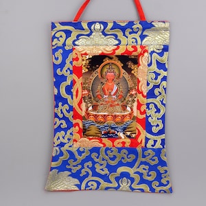May include: A colorful Tibetan Buddhist thangka depicting a deity, likely a bodhisattva, seated on a lotus throne. The thangka is framed with a red and gold border and is surrounded by a blue and gold patterned fabric.