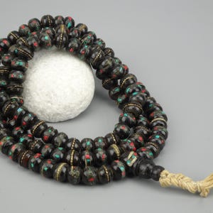 Könnte beinhalten: Eine schwarze Mala-Gebetskette mit roten und grünen Akzenten. Die Perlen sind aus Holz und haben einen goldenen Rand. Die Kette ist auf einer Schnur mit einem Quasten befestigt.